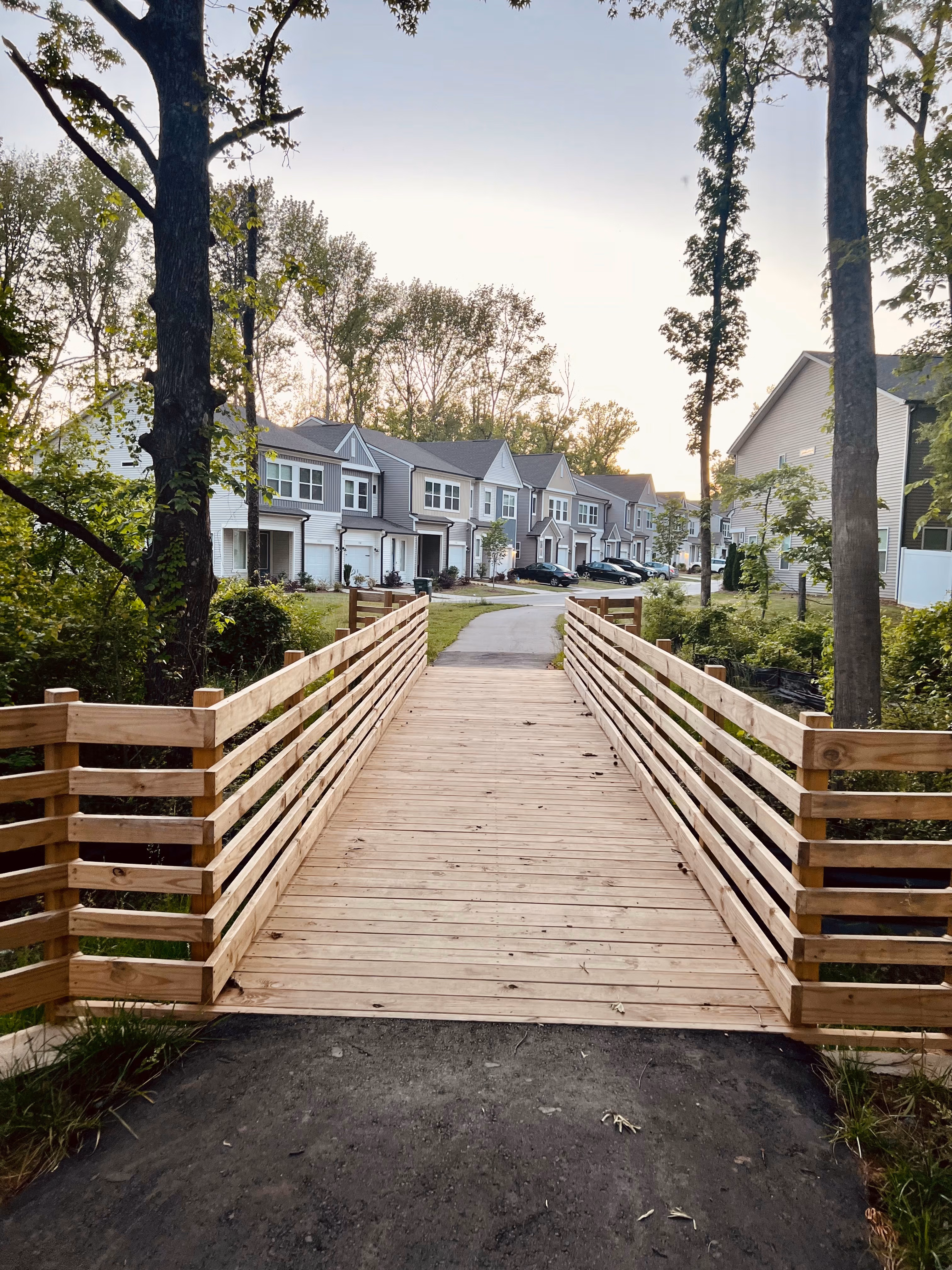 A wooden walkway leads across a grassy area towards a row of residential buildings surrounded by trees in the background.