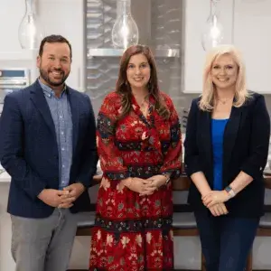 The image shows three people standing together in what appears to be a professional or business setting, with a background of shelves and lighting fixtures.